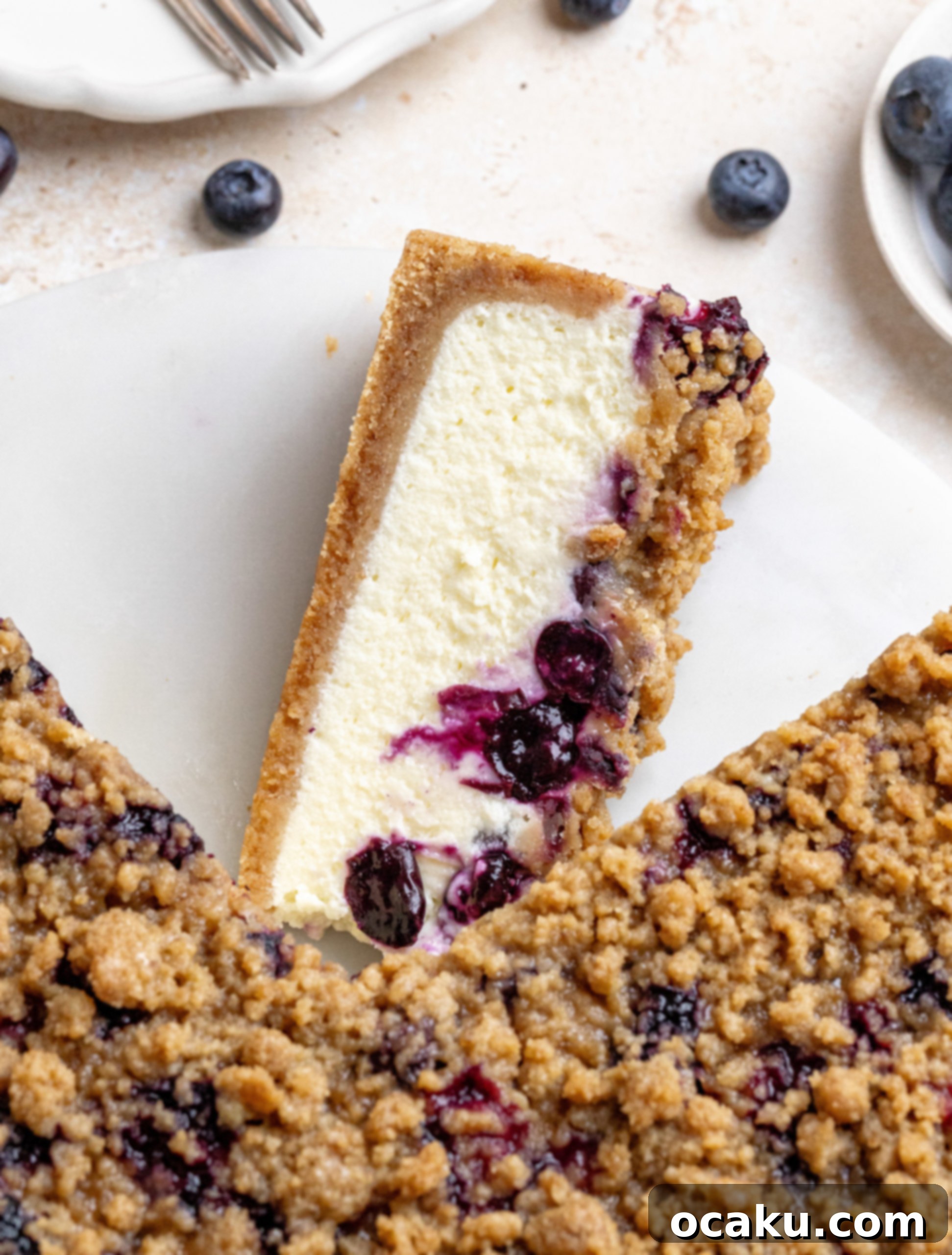 A slice of blueberry crumble cheesecake on a plate, showing layers of crust, creamy filling, blueberries, and crumble topping.