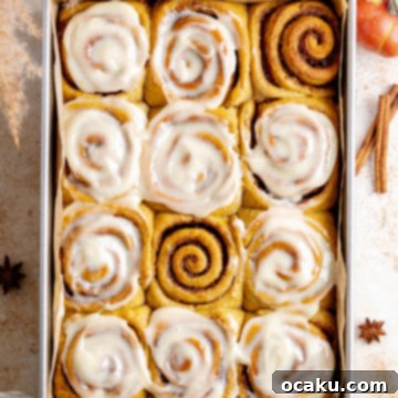 Freshly baked pumpkin cinnamon rolls in a square baking dish, topped with generous maple cream cheese frosting.