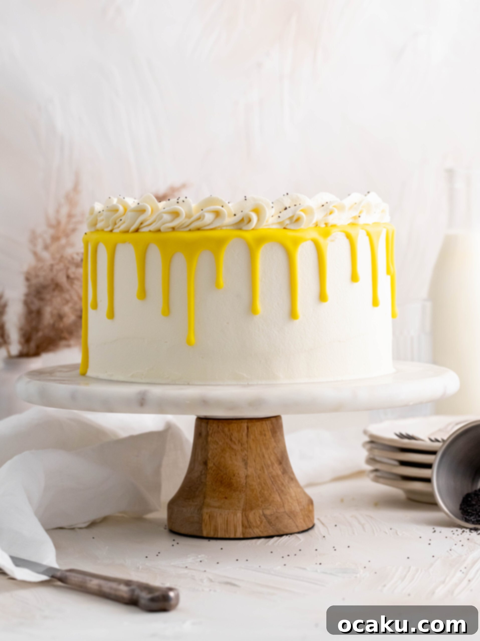 A stunning lemon poppy seed layer cake adorned with a white chocolate drip and buttercream wreath, presented on a elegant cake stand.