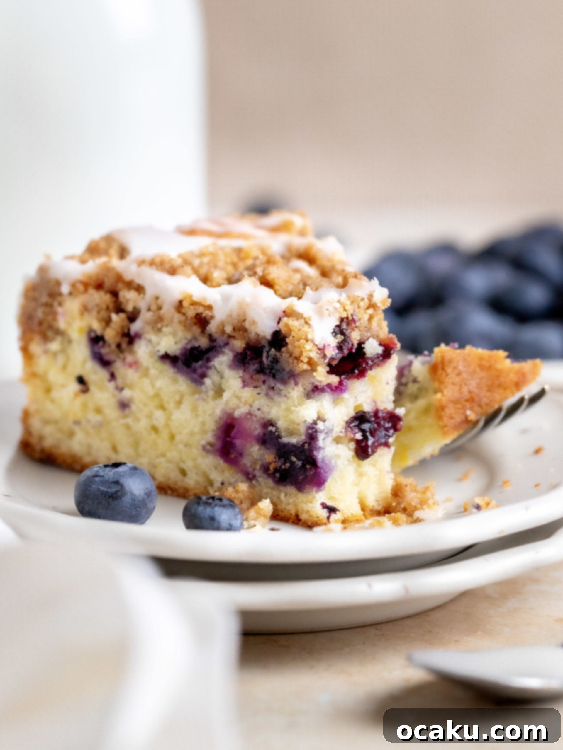 A delicious slice of homemade blueberry coffee cake with crumb topping and lemon glaze, served on a white plate.