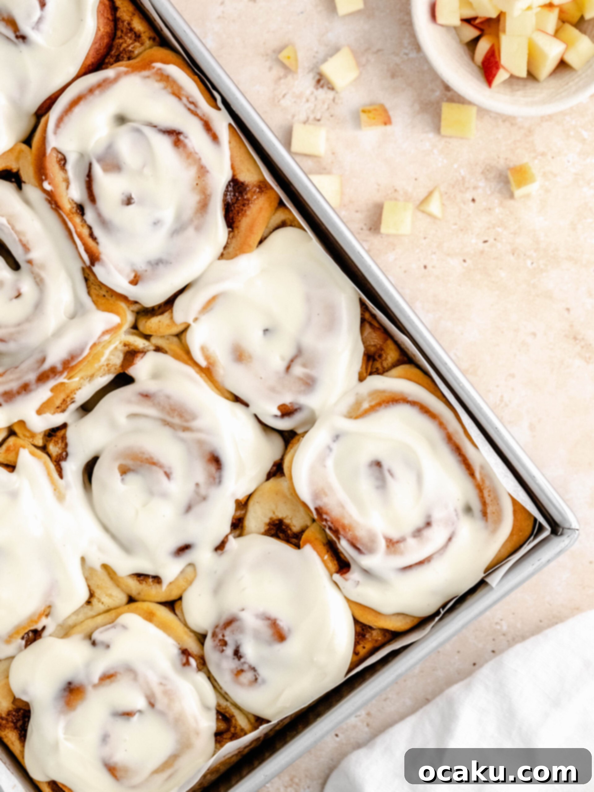 Freshly baked cinnamon rolls with apple pie filling in a baking pan, topped with cream cheese frosting.