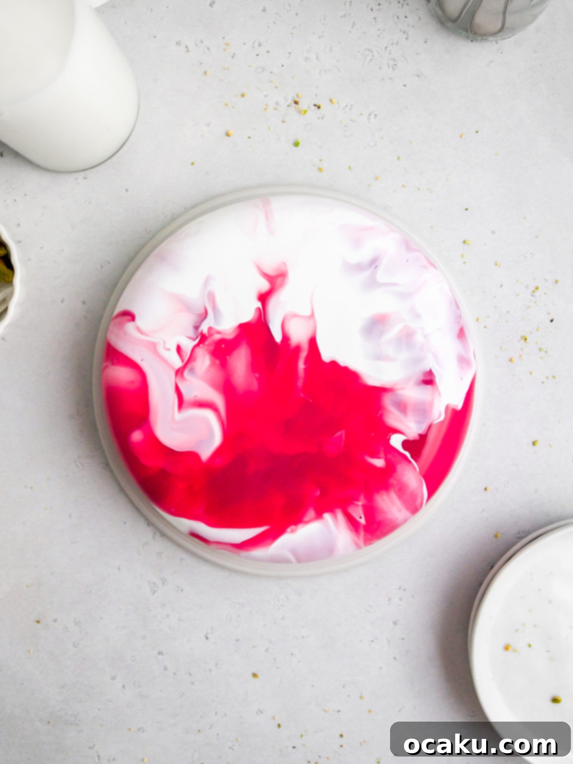 A beautifully crafted strawberry and pistachio mousse cake, topped with a shimmering mirror glaze, presented on a white plate with fresh strawberries and pistachios for garnish.