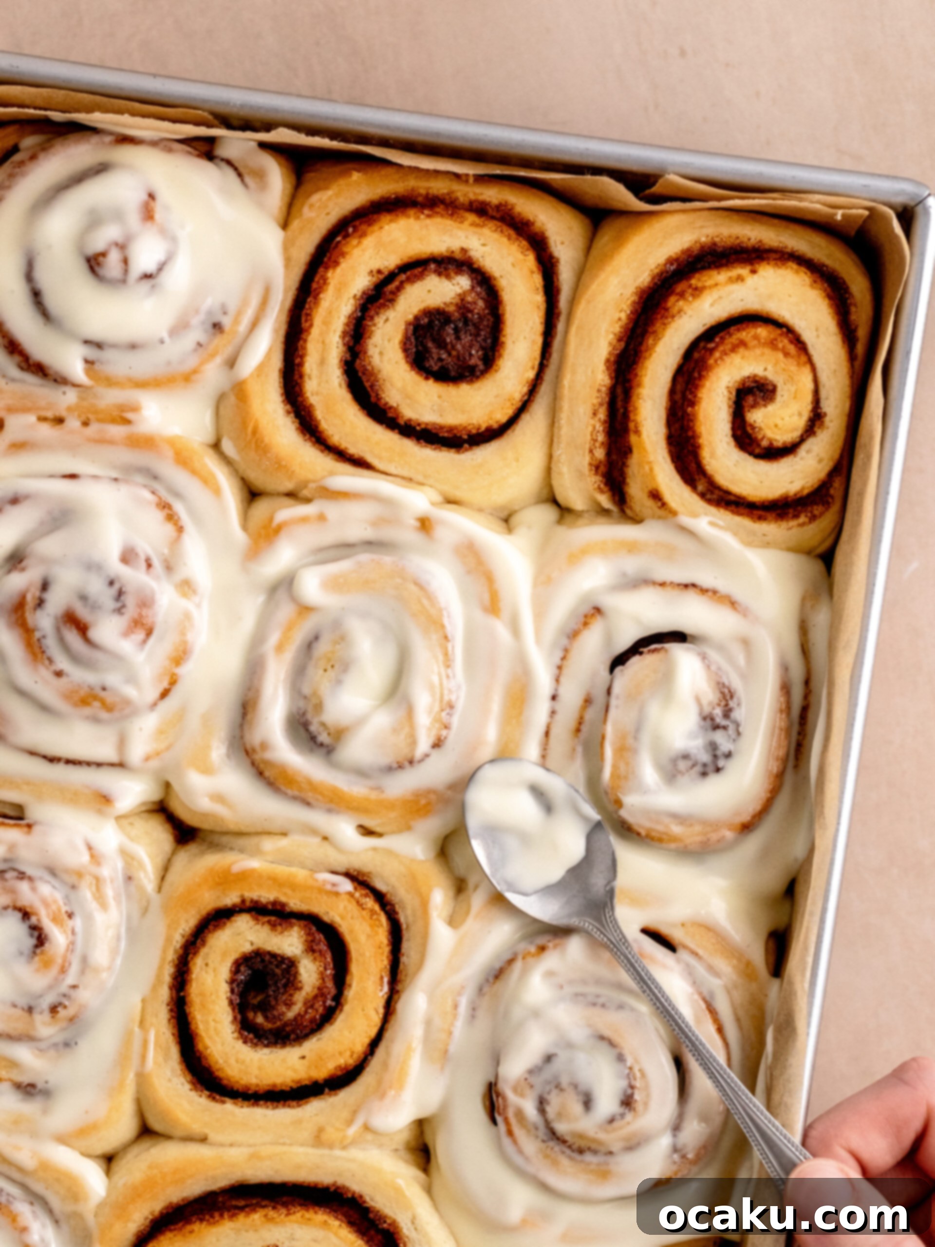 Cream cheese frosting being spread smoothly over the warm brioche cinnamon rolls