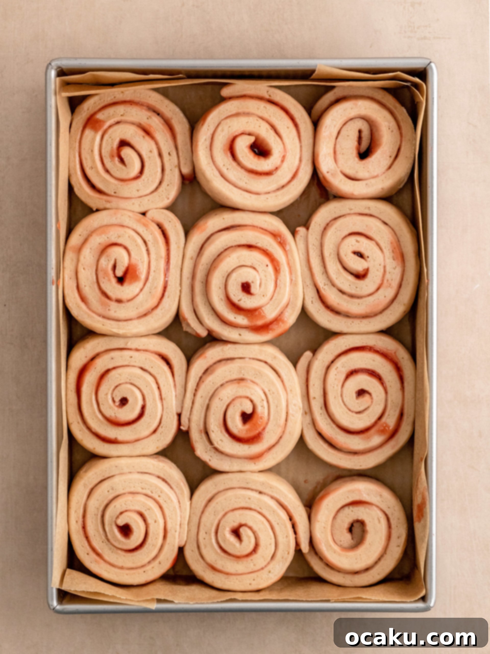 A hand spreading a generous layer of pink strawberry cream cheese frosting over warm, baked strawberry rolls.