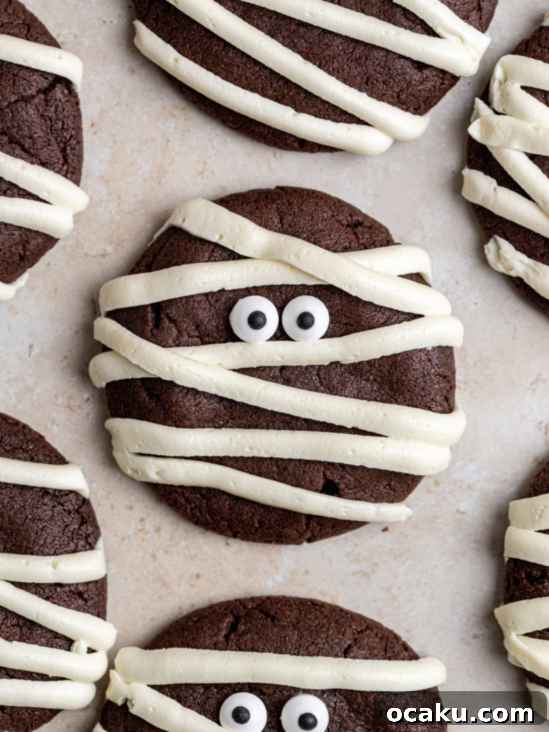 Spooky Halloween Mummy Cookies 2 Close-up of three Halloween Mummy Cookies with fudgy chocolate base and vanilla buttercream wrapping, decorated with candy eyes on a white surface.
